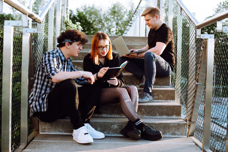 Concentrated small group of people, two men and woman in glasses use laptop, book on stairs outdoors. Business and studyの写真素材