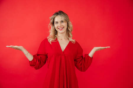 Portrait of attractive young woman in bright red dress spreading arms to sides isolated on red background. Showing result, advertisement, beautiful smile. Fashion and beauty industryの写真素材