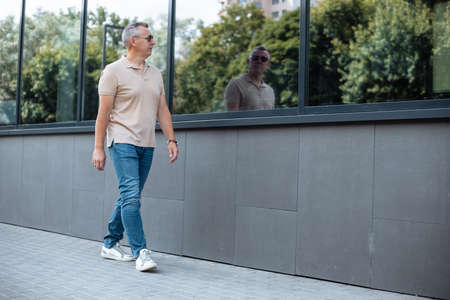 Portrait side view from below of old man in sunglasses walking on street along office building and looking on silhouette reflection in window. Urban lifestyle, outdoor recreation, summer leisureの写真素材