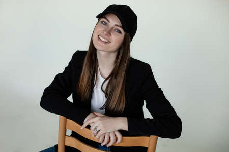 Smiling young girl sitting on chair, gray studio background, copy space. Female casual black blazer...