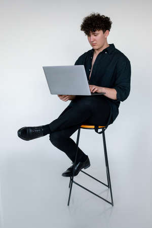 Full length photo of upset young curly guy in total black look sitting cross legged on chair and playing computer games on modern laptop, gray studio background. Modern Internet realityの写真素材