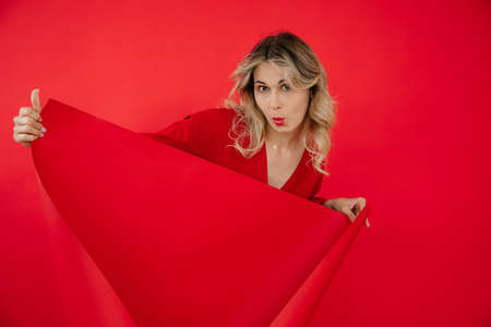 Portrait of cheerful cute woman in red outfit uncovering red empty whatman paper isolated on red background. Marketing and advertisement, free copy space. Fashion and beauty industryの写真素材