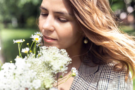 Portrait closeup of young charming woman smelling and enjoying aroma of bouquet plucked white wild flowers. Walking in park on sunny day, outdoor relaxation, beauty of nature. Allergy free peopleの写真素材