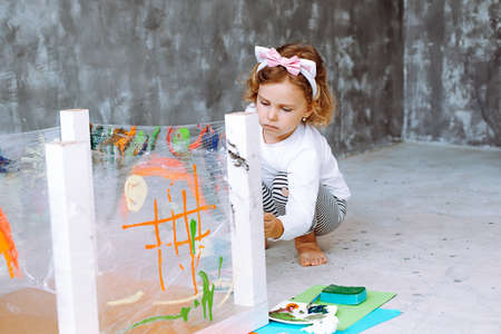Portrait of serious barefoot little girl sit on haunches on art lesson, painting drawing with brush on stretch film.の写真素材