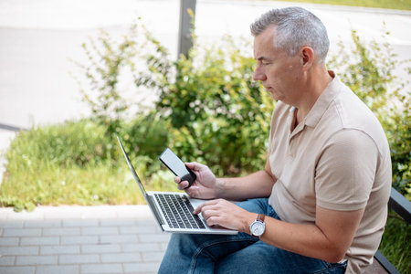 Aged gray haired businessman sitting on bench in green city park with smartphone and typing on digital laptop. Modern lifestyle, remote job, social communication. Technology and people conceptの写真素材