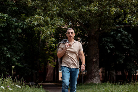 Old brutal man in black sunglasses walking along path in city forest park on sunny day. Urban lifestyle, free time, vacation, outdoor recreation, leisure. Nature and people conceptの写真素材