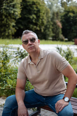 Portrait of old brutal man in black sunglasses sitting on bench in green city square and resting on sunny day. Urban lifestyle, free time, outdoor recreation, leisure. Nature and people conceptの写真素材