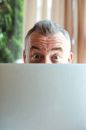 Closeup photo of cheerful mature businessman working with laptop computer and peeking out from behind monitor, free copy space. Remote job, working time, freelance, busy people, having funの写真素材