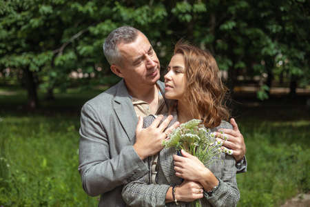 Portrait of loving couple gently hugging in green park on sunny day, nature background. Adult man and young woman walking with bouquet of wild flowers outdoors. Romantic date, true love, feelingsの写真素材
