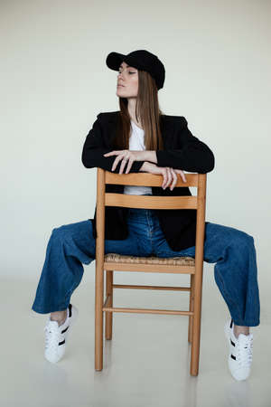 Full length photo of attractive girl sitting on chair, gray studio background, copy space. Female casual black blazer and cap, jeans and gumshoes. Fashionable women clothing, headdresses and footwearの写真素材