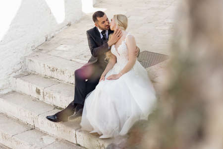 Beautiful couple sitting on old white concrete stairs. Young blond woman bride in white dress kissing man groom in suit.の写真素材