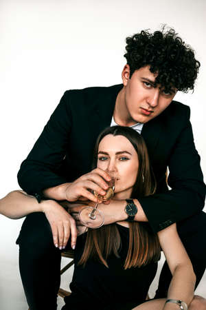 Portrait of cool couple in black outfits celebrating holiday with glass of champagne, gray studio background, copy space. Brutal guy sitting and embracing beautiful girlfriend. Couples style conceptの写真素材