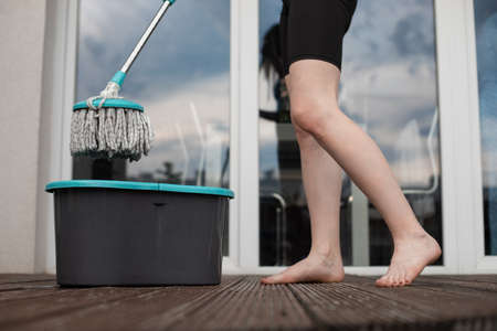 Cropped photo of young barefoot woman holding spin mop over grey plastic bucket, cleaning wooden...