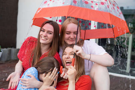 Group of young laughing stunning women sitting on wooden veranda, hiding from rain under umbrella with dots, having fun.の写真素材