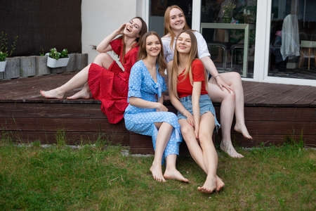 Group of young smiling gorgeous barefoot women sitting on wooden veranda near green grass, looking at camera, laughing.の写真素材
