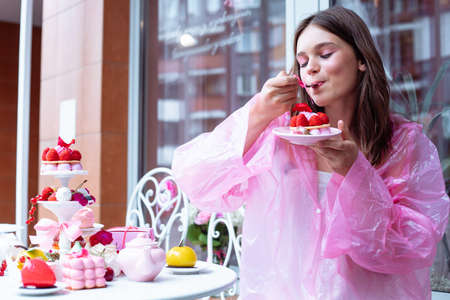 Pretty teenage girl in pink raincoat joying, tasting sweet berry dessert cakes from the table in cafe. Birthday eventの写真素材