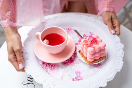 Sugar pink cube ball velours mousse cake dessert on plate with cup of tea in cropped woman hands....