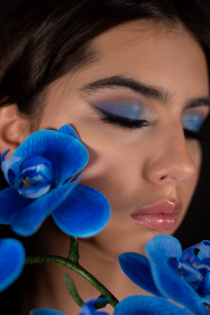 Close-up of young woman with closed eyes, bright blue eyeshadows, pink lips holding near face branch of blue orchid.の写真素材