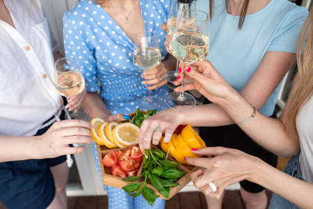 Cropped photo of young women holding cut vegetables, clinking glasses of white wine, standing near house. Top view.の写真素材