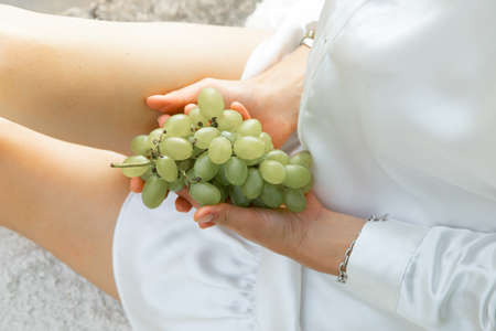 Cropped photo of young woman wearing white dress, sitting, holding delicious bunch of fresh ripe green grapes in hands.の写真素材