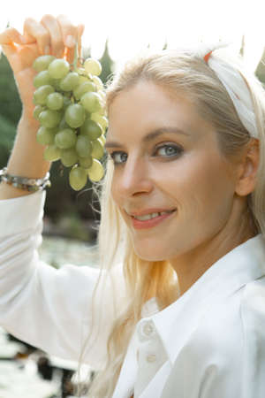 Portrait of young perfect smiling woman wearing white dress, sitting, raising bunch of green grapes in hand at face.の写真素材