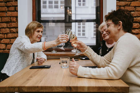 Three senior women friends having dinner at restaurant, attractive lady spending time together indoors. old friends drinking wine in modern apartment near windowの写真素材