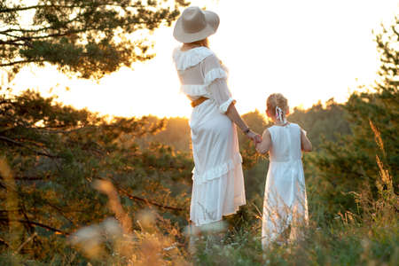 Back view of wonderful family wearing white dresses, standing near pines in park forest looking at sunset in summer.の写真素材