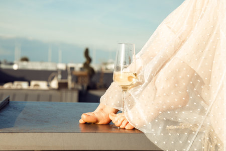 Close-up portrait of cropped barefoot woman feet in white transparent dress near wineglass with drink champagne on roofの写真素材