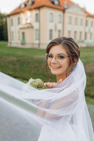 Joyful, cheerful, smiling young bride in glasses in white elegant dress covered by veil with bouquet against new houseの写真素材