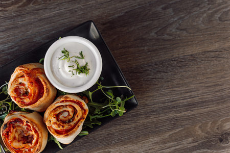 Appetizing pepperoni pastry rolls with filling, greenery and sauce on black elegant plate on wooden table in restaurantの写真素材