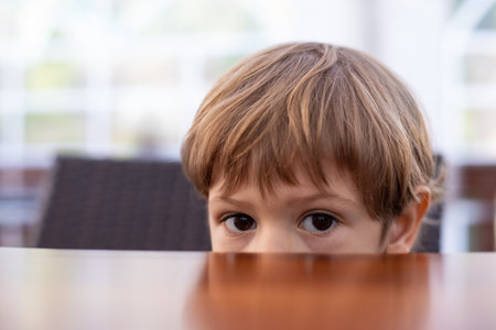 Fearful little boy hide half face under wooden table in cafe hall closeup, free copy space. Pretty kid of kindergarten age scared to sit in kitchen. Morning, breakfast, phobia, aloneの写真素材