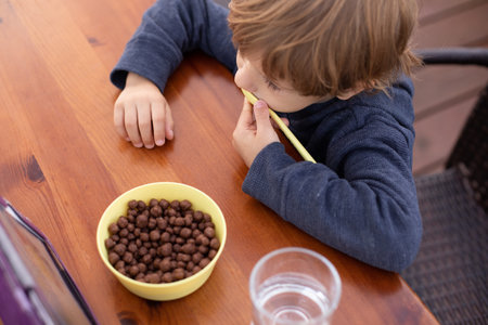 Little boy sit on chair at table and watch cartoon on tablet computer top view. Kid of kindergarten age eat chocolate balls with milk and drink water. Ready breakfast, gadget addictionの写真素材