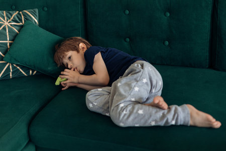 Portrait of upset, sad and depressed barefoot little boy lying on green sofa, sucking thumb finger. Psychological stressの写真素材
