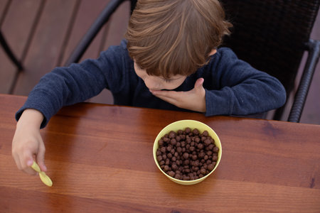 Little boy sitting on chair at brown wooden table in kitchen and having breakfast with chocolate balls top view. Kid of kindergarten age trifle spoon in hand. Ready breakfast, not hungry childの写真素材