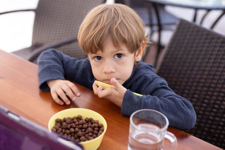 Unhappy little boy sit at table and watch cartoon on tablet computer top view. Kid of kindergarten age eat chocolate balls with milk and drink water. Autism, mental disease, deviation, health problemの写真素材