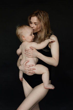 Vertical portrait of young blonde woman, mother holding and carrying baby in arms, kissing and embracing in black studioの写真素材
