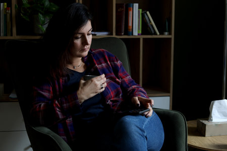 Concentrated, serious brunette businesswoman in plaid shirt use mobile phone and drink coffee in...