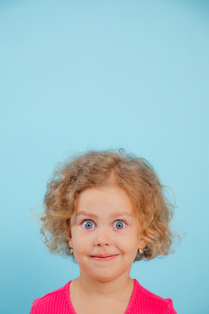 Portrait of smiling little girl with curly hair, wide-opened blue eyes wearing pink T-shirt, looking...