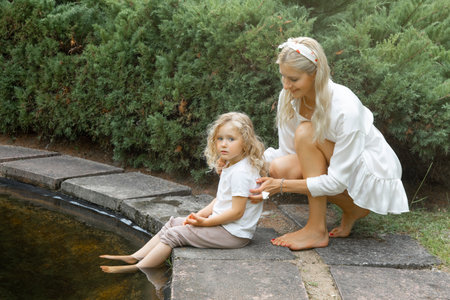 Portrait of little girl sitting on edge of pond with feet in water in park, while young woman creeping up to daughter.の写真素材