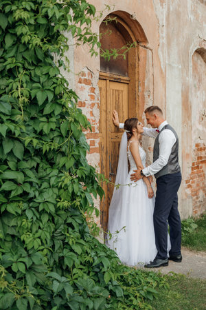 Side view of happy wedding couple stand outside near old building ivy-covered in summer. Young man groom hugging woman.の写真素材