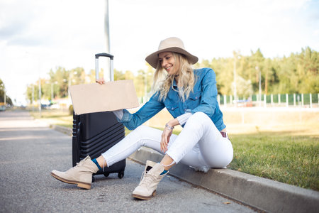 Portrait of young happy woman holding blank cardboard, sitting on road near black suitcase, green grass. Travelling.の写真素材
