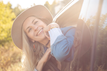 Dreaming woman joyfully leave city sticking head out of open car window. Young blonde lady in hat with happy mood go to auto trip. Travelling, escapism, vacations, active lifestyleの写真素材