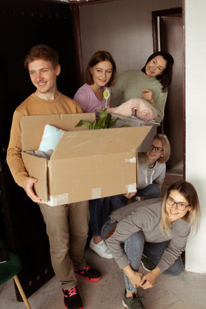 Vertical curious helpful energetic teenagers carrying carton boxes with plants, clothes near door. First grade educationの写真素材