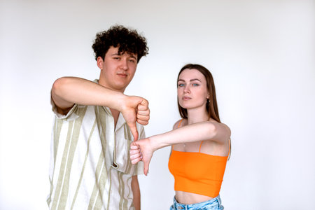 Portrait of young serious couple posing on white background. Young curly confident man, woman showing thumbs down sign.の写真素材