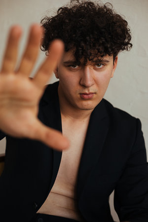 Portrait of young curly serious gorgeous man wearing black jacket, sitting, stretching hand forward with open palm.の写真素材