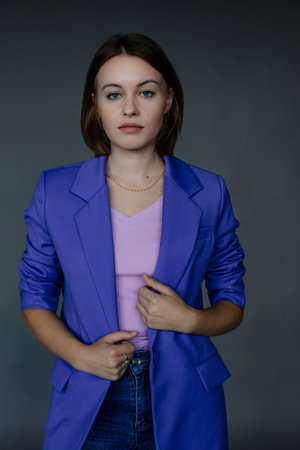 Portrait of young serious beautiful blue-eyed businesswoman holding edges of blue jacket, posing on grey background.の写真素材