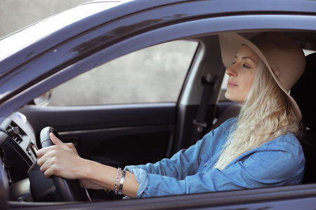 Young woman with long fair hair in denim shirt, beige floppy hat, sitting in black car, holding...