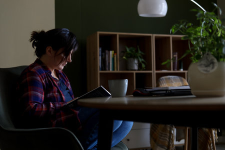 Tranquil, silent, depressed black haired mature woman read book in armchair in dark room. Frustration, emotional stressの写真素材