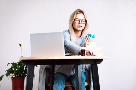 Portrait of young woman sitting near laptop, holding bottles with blue caps, taking pills blister, showing medicine.の写真素材