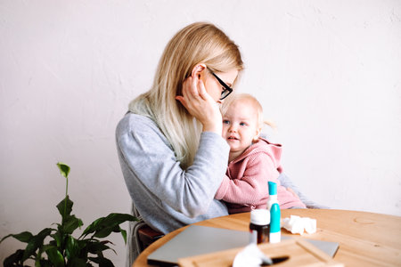 Side view of young mother woman sitting at table, holding little sad baby girl, calming down child at home. Sick time.の写真素材
